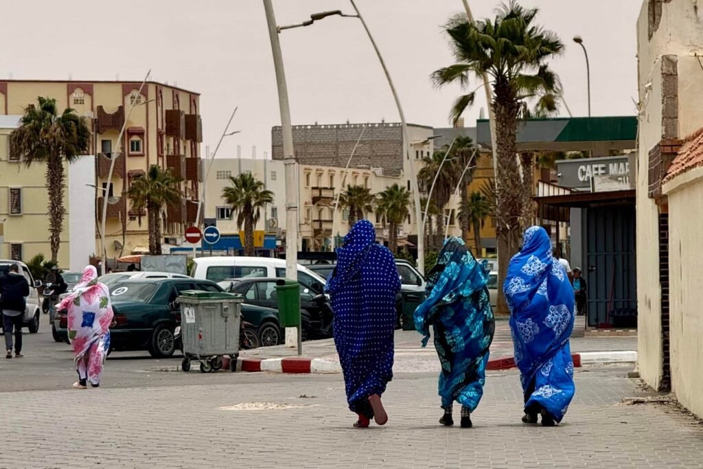Cuatro mujeres saharauis de espaldas caminando por una calle de Boujdour, vestidas con coloridas melhfas azules y rosas, entre edificios y palmeras